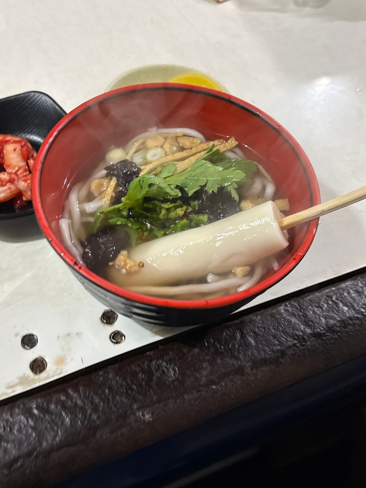 붉은 그릇에 담긴 우동과 그 위에 올린 꼬치 떡, 오뎅