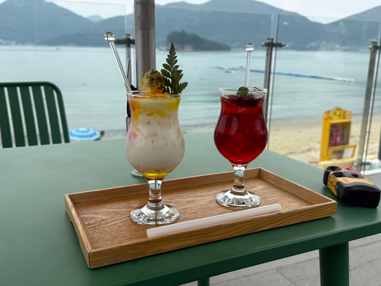 A white drink and a red drink on a wooden tray on a green table, with the sea behind them