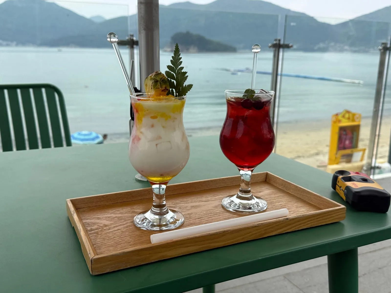 A white drink and a red drink on a wooden tray on a green table, with the sea behind them