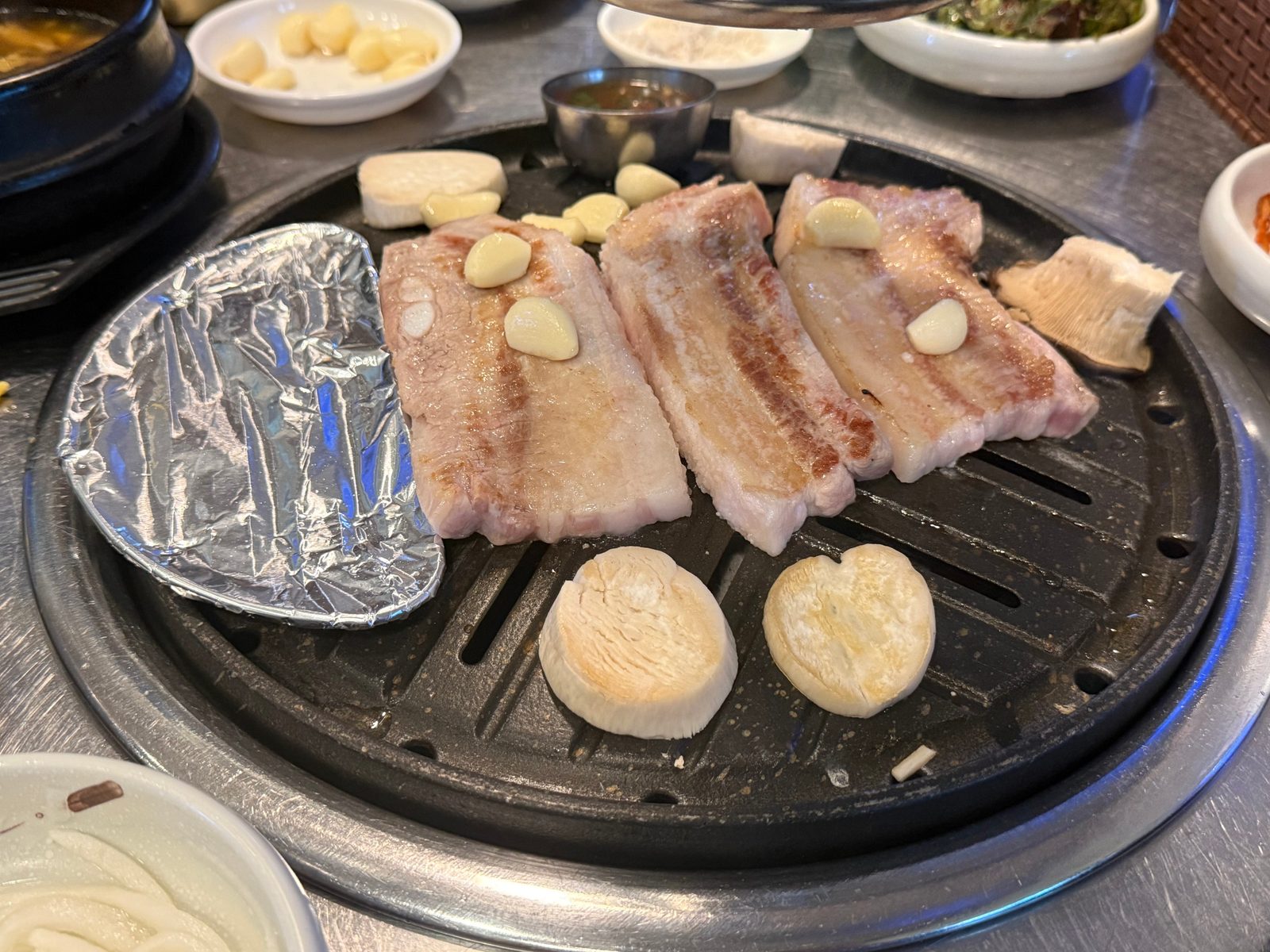 Three thick slabs of pork belly on a round grill plate with garlic and foil