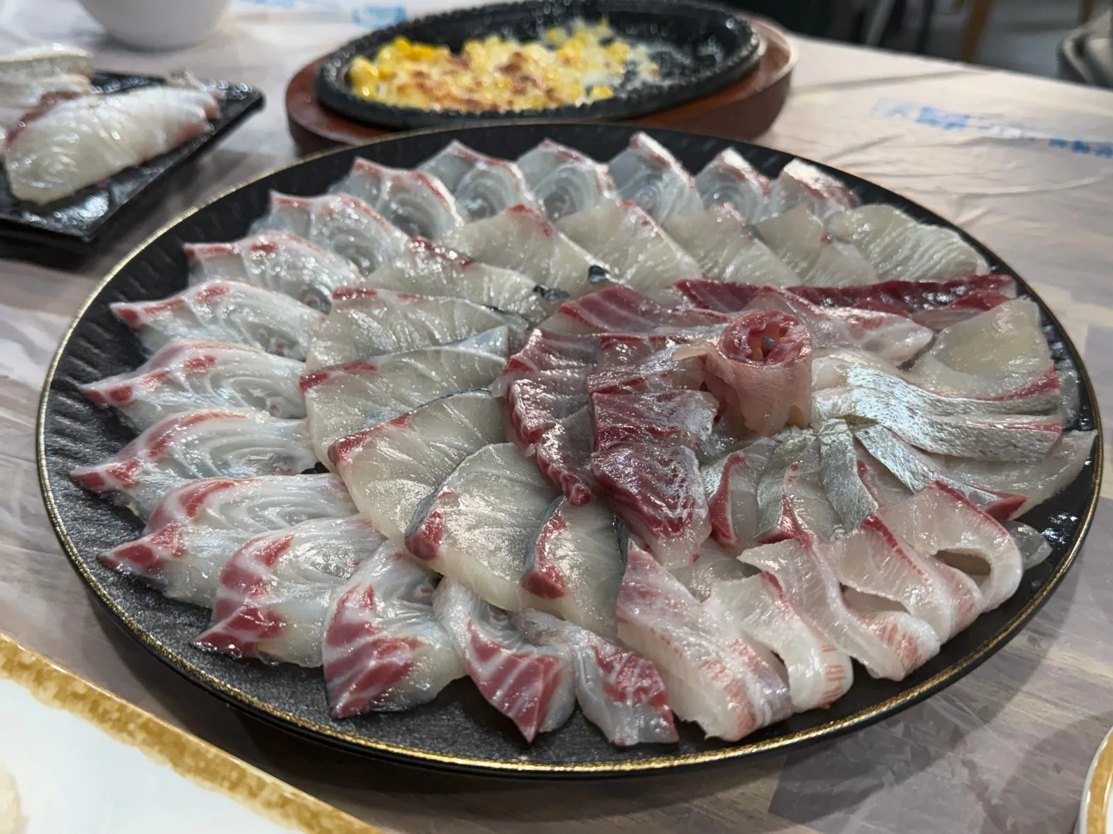 Sashimi arranged in a circle like petals on a black plate, with corn cheese behind