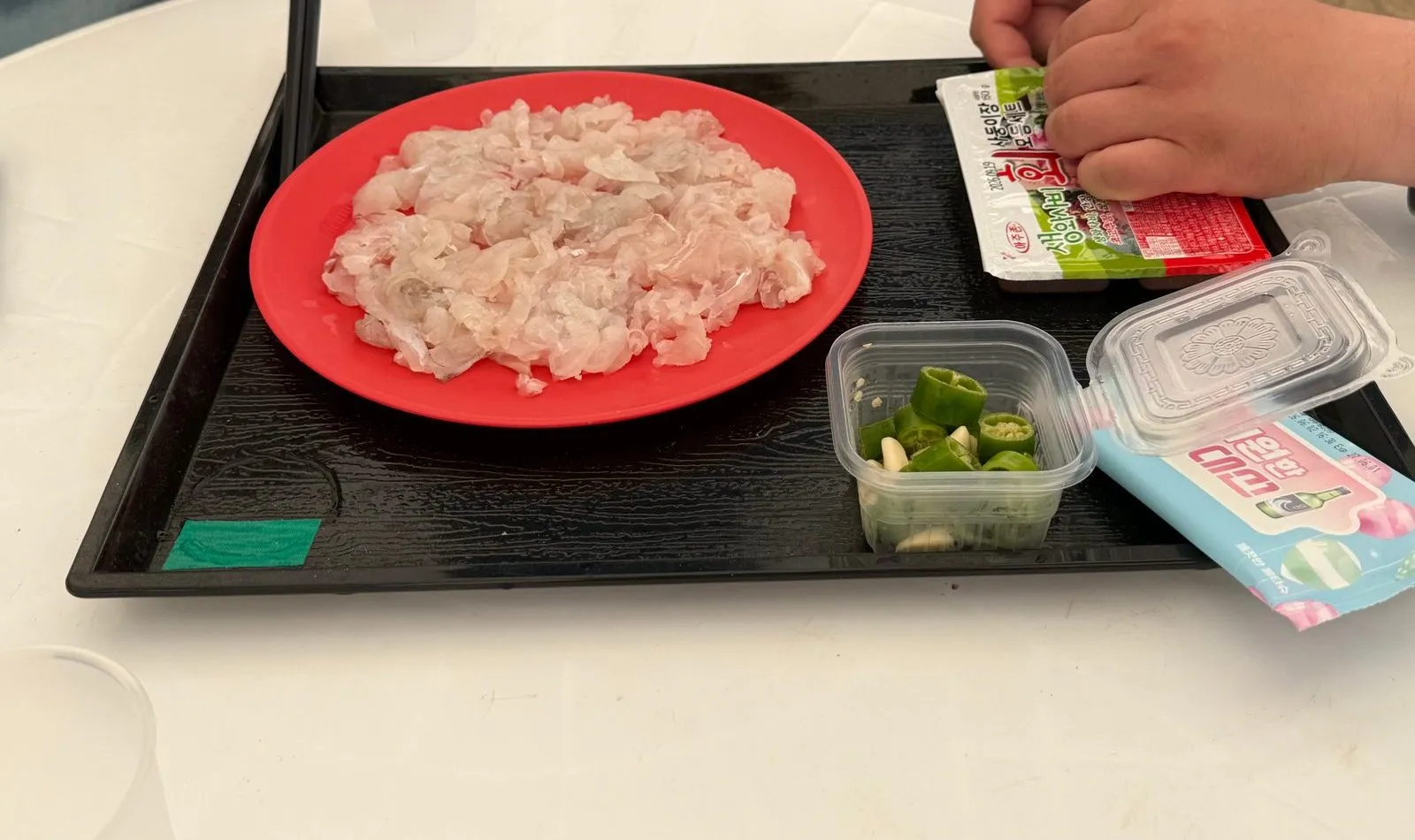 A tray with sliced raw fish on a red plate, chili sauce packets, and green chilies