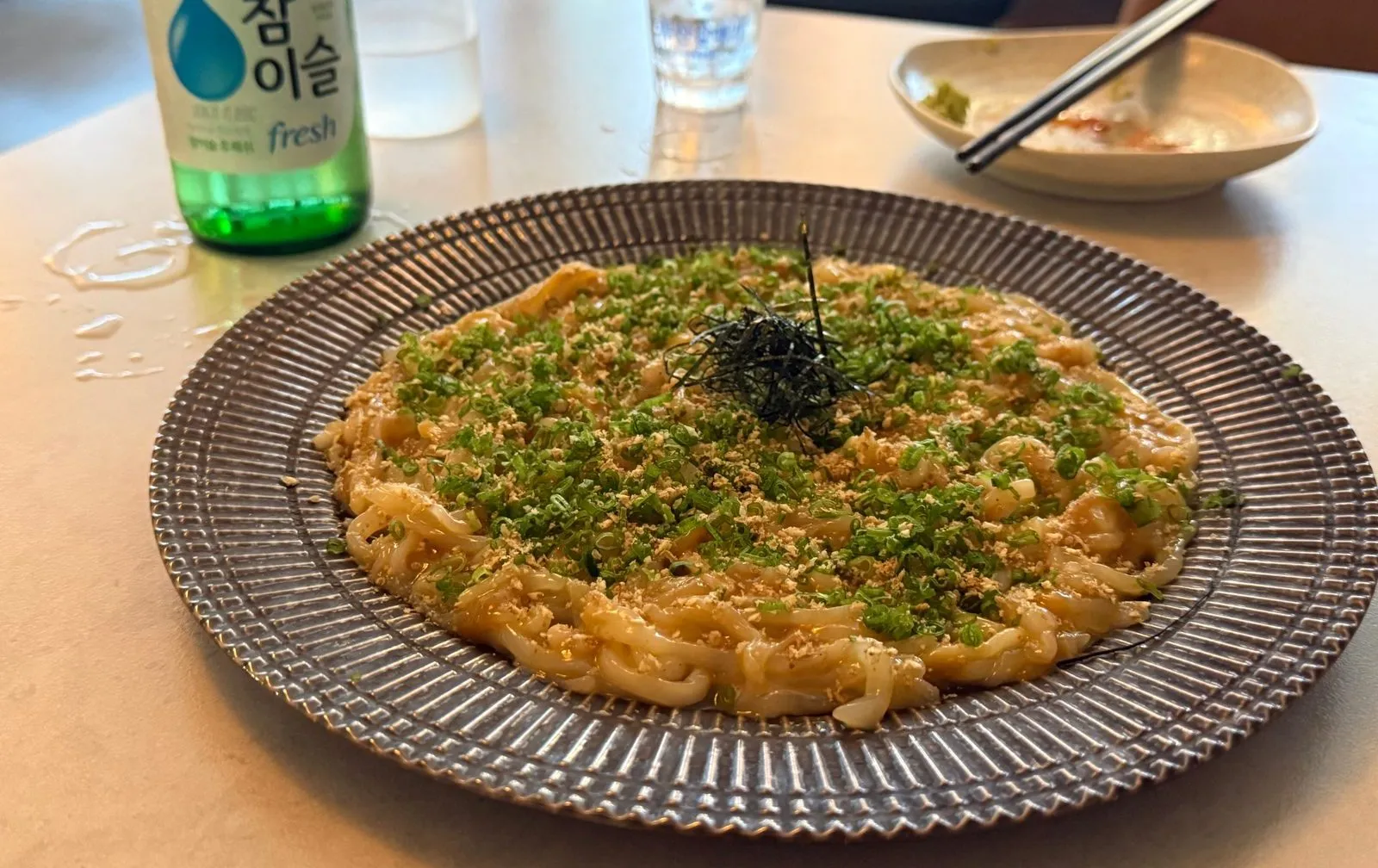A plate of thinly cut squid topped with chopped green onion, with a soju bottle behind