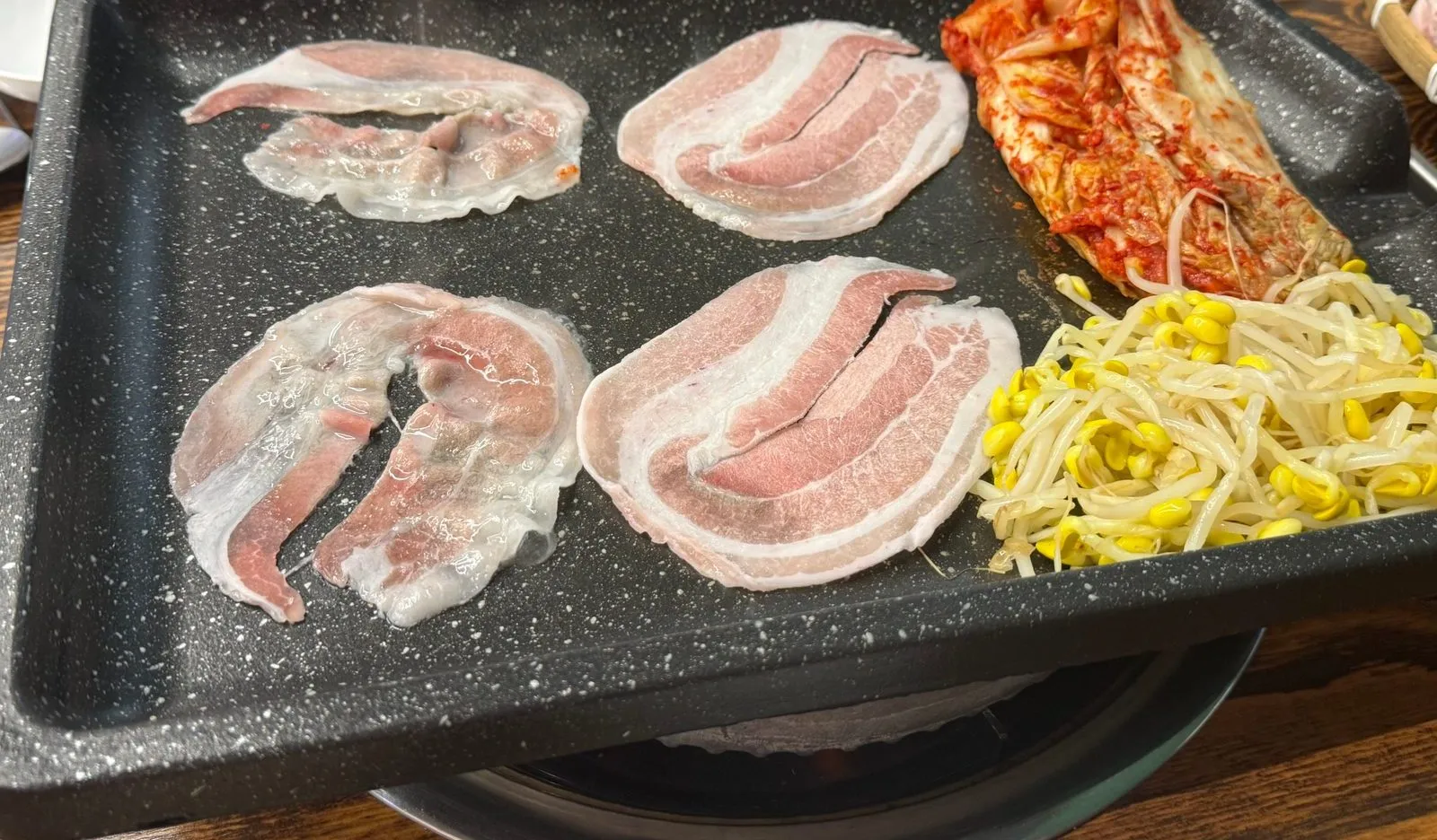 Pork belly slices laid out one by one on a grill plate with kimchi and bean sprouts