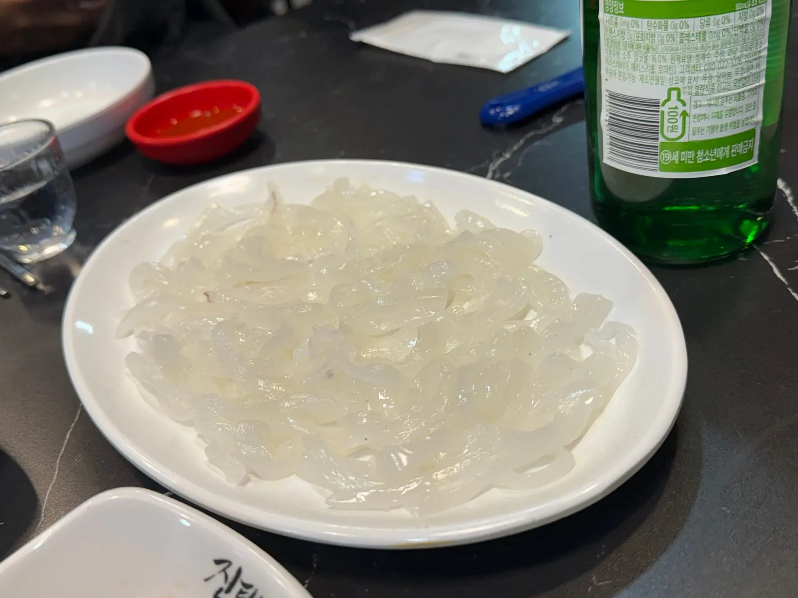 Thinly sliced raw squid on a white plate, with a soju bottle beside it