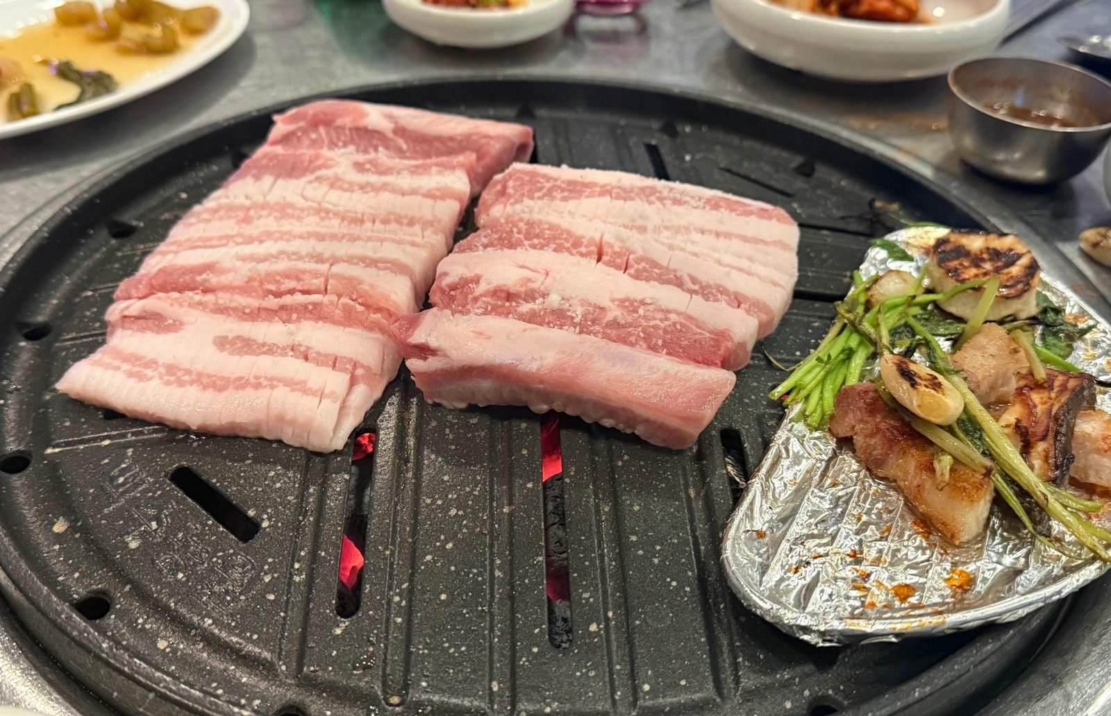 숯불 불판 위에 올린 두툼한 삼겹살 두 덩이와 은박지에 덜어 둔 구운 고기